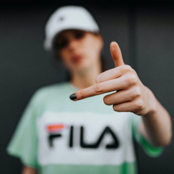 Young person in a Fila t-shirt and cap pointing at the camera with a focused expression, leader of the haters, with the background out of focus.