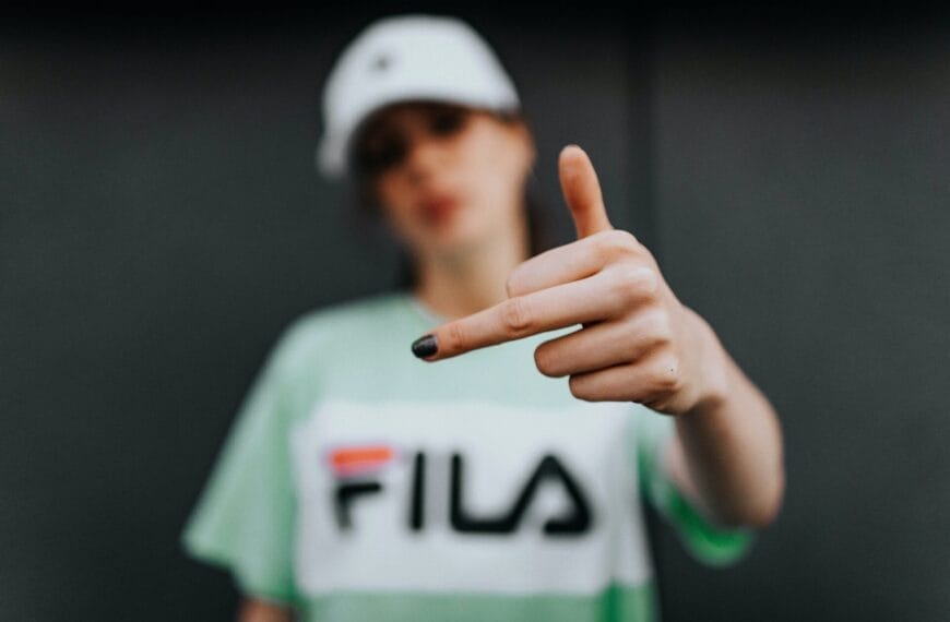 Young person in a Fila t-shirt and cap pointing at the camera with a focused expression, leader of the haters, with the background out of focus.