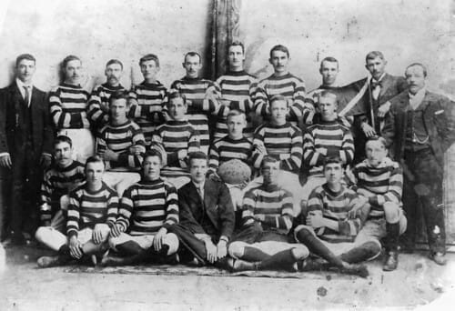 Historical black and white photo of a football team in striped uniforms, posing with coaches and a ball, circa early 20th century.
