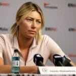 A person at a press conference table, surrounded by microphones and wearing a light-colored shirt, looks thoughtful while addressing controversial sports issues.