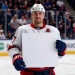 A hockey player, possibly pondering NHL player contracts, stands on the ice holding a blank white sign, with a lively crowd in the backdrop.