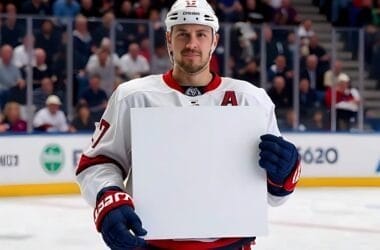 A hockey player, possibly pondering NHL player contracts, stands on the ice holding a blank white sign, with a lively crowd in the backdrop.