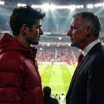 Two men in a stadium, facing each other with polarizing sports views, one in a red jacket and the other in a suit, with a soccer field in the background.