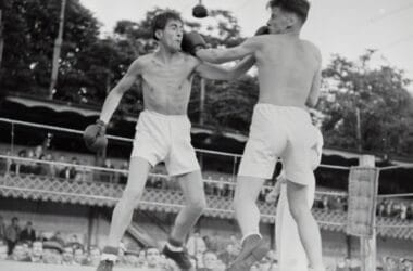 Two boxers face off in the ring, with one landing a punch on the other's face as spectators cheer from the background. The atmosphere buzzes with hot sports takes, fueling the electrifying energy of the match.