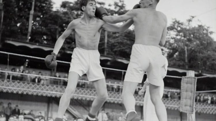 Two boxers face off in the ring, with one landing a punch on the other's face as spectators cheer from the background. The atmosphere buzzes with hot sports takes, fueling the electrifying energy of the match.