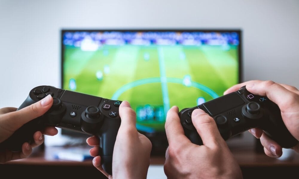 Two people holding game controllers are engrossed in sports video content, as a soccer video game unfolds on the TV screen.