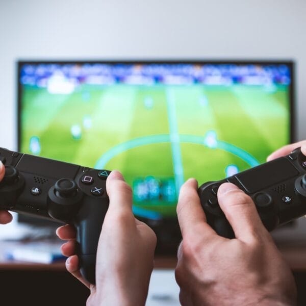 Two people holding game controllers are engrossed in sports video content, as a soccer video game unfolds on the TV screen.