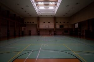 A dimly lit indoor sports hall subtly highlights the basketball hoop and court lines, inviting players to explore the subjectivity in basketball, where every angle can shift perception.