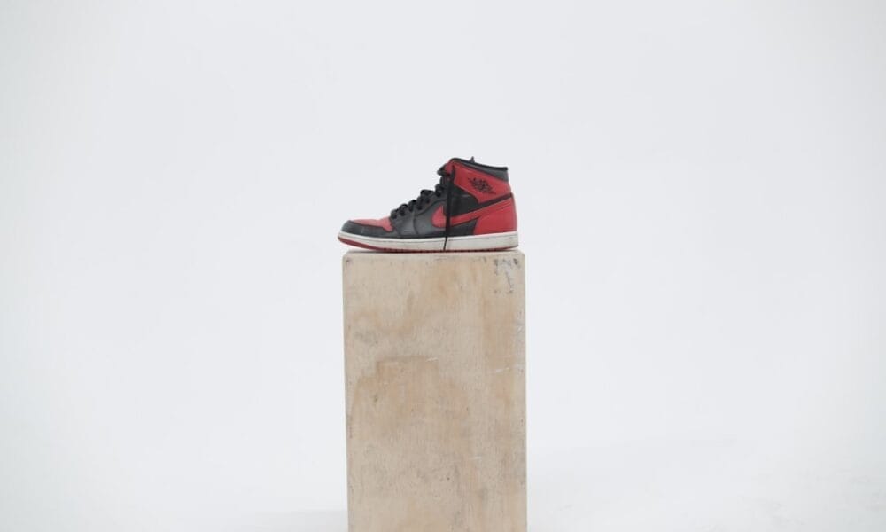 A red and black high-top sneaker reminiscent of 90s basketball fashion is displayed on a wooden box against a plain white background.