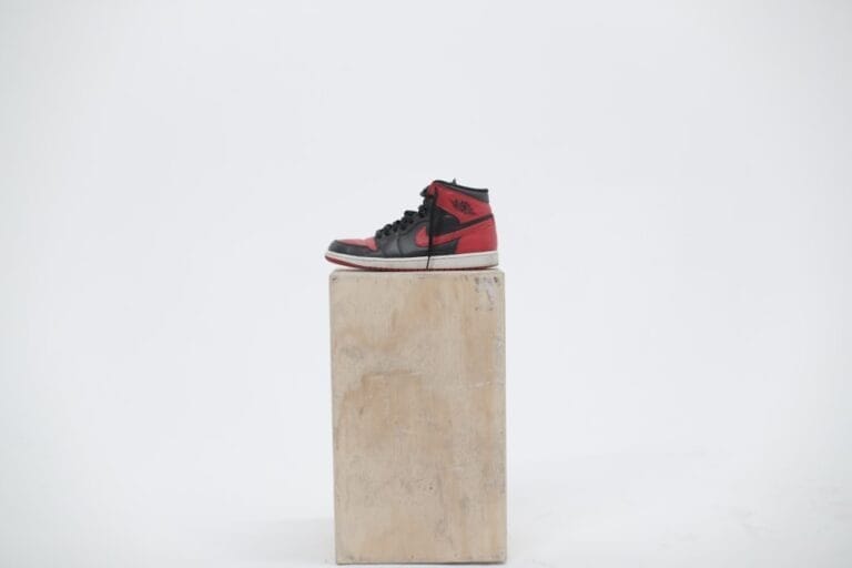 A red and black high-top sneaker reminiscent of 90s basketball fashion is displayed on a wooden box against a plain white background.