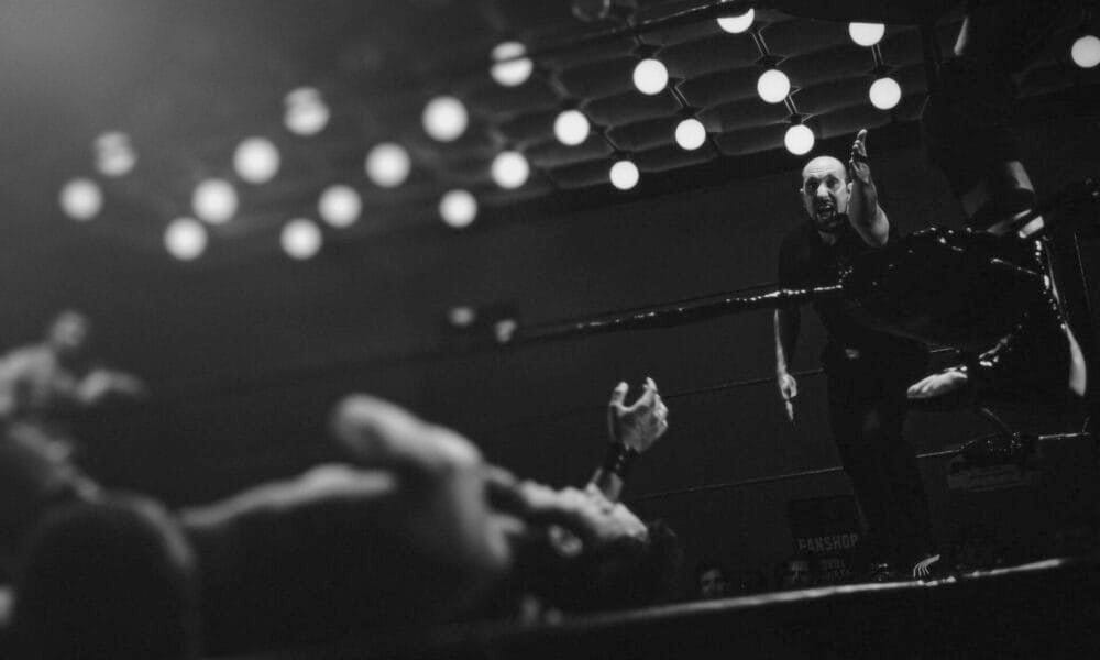 A wrestling referee points as a wrestler lies on the mat, surrounded by bright ceiling lights.