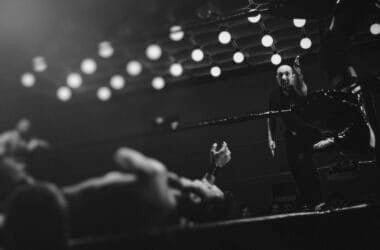 A wrestling referee points as a wrestler lies on the mat, surrounded by bright ceiling lights.