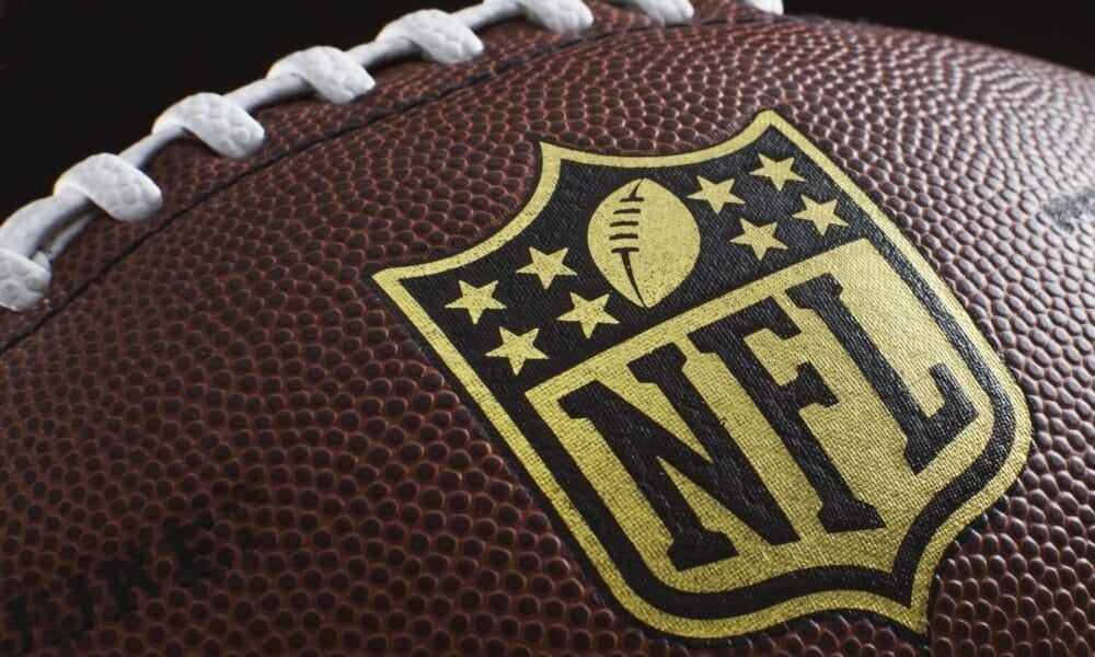 Close-up of an American football showcasing the NFL logo, a symbol of rich NFL traditions. The brown leather surface features white laces and textured pebbling, with the logo prominently centered and highlighted against the football.