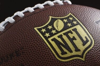 Close-up of an American football showcasing the NFL logo, a symbol of rich NFL traditions. The brown leather surface features white laces and textured pebbling, with the logo prominently centered and highlighted against the football.