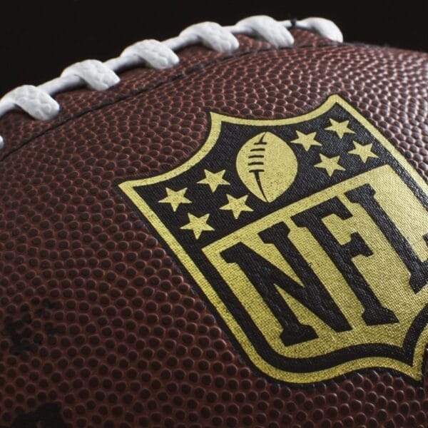 Close-up of an American football showcasing the NFL logo, a symbol of rich NFL traditions. The brown leather surface features white laces and textured pebbling, with the logo prominently centered and highlighted against the football.