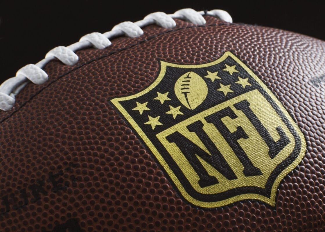 Close-up of an American football showcasing the NFL logo, a symbol of rich NFL traditions. The brown leather surface features white laces and textured pebbling, with the logo prominently centered and highlighted against the football.