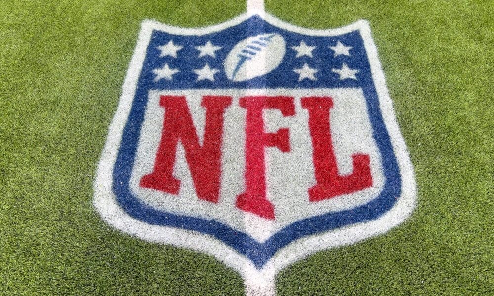 The image captures the NFL logo, a symbol of tradition, painted on a grassy field. The design features a shield shape with a football at the top, encircled by stars, and bold red "NFL" letters at the center. A white boundary line gracefully crosses through this iconic emblem.