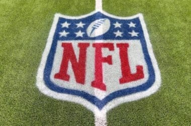 The image captures the NFL logo, a symbol of tradition, painted on a grassy field. The design features a shield shape with a football at the top, encircled by stars, and bold red "NFL" letters at the center. A white boundary line gracefully crosses through this iconic emblem.