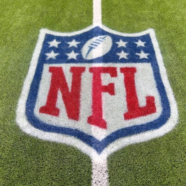 The image captures the NFL logo, a symbol of tradition, painted on a grassy field. The design features a shield shape with a football at the top, encircled by stars, and bold red "NFL" letters at the center. A white boundary line gracefully crosses through this iconic emblem.