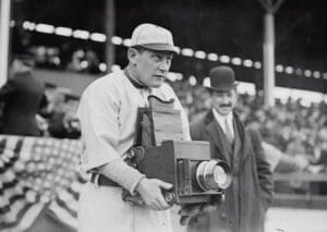 A baseball player intrigued by sabermetrics holds a vintage camera at the stadium, while a man in a suit and bowler hat stands nearby.