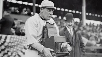 A baseball player intrigued by sabermetrics holds a vintage camera at the stadium, while a man in a suit and bowler hat stands nearby.