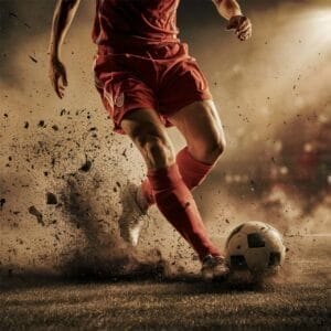 A soccer player in a red uniform kicks a ball on a dirt field, with soil flying around—a scene that might soon be part of the future of sports as traditional fields give way to innovative playing environments.