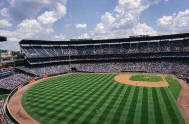 A wide-angle view of a baseball stadium captures an MLB tradition in progress. The field, boasting alternating green stripes, is impeccably maintained. Sparse crowds enjoy the game under a partly cloudy sky, adding to the timeless atmosphere of America's favorite pastime.