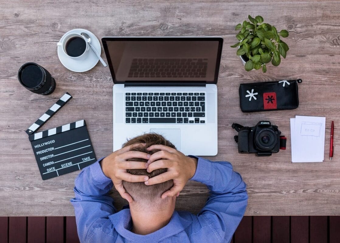 A person sits at a desk, hands on head in thought, surrounded by a laptop, camera, clapperboard, and the tools of sports media coverage—coffee and a potted plant completing the scene.