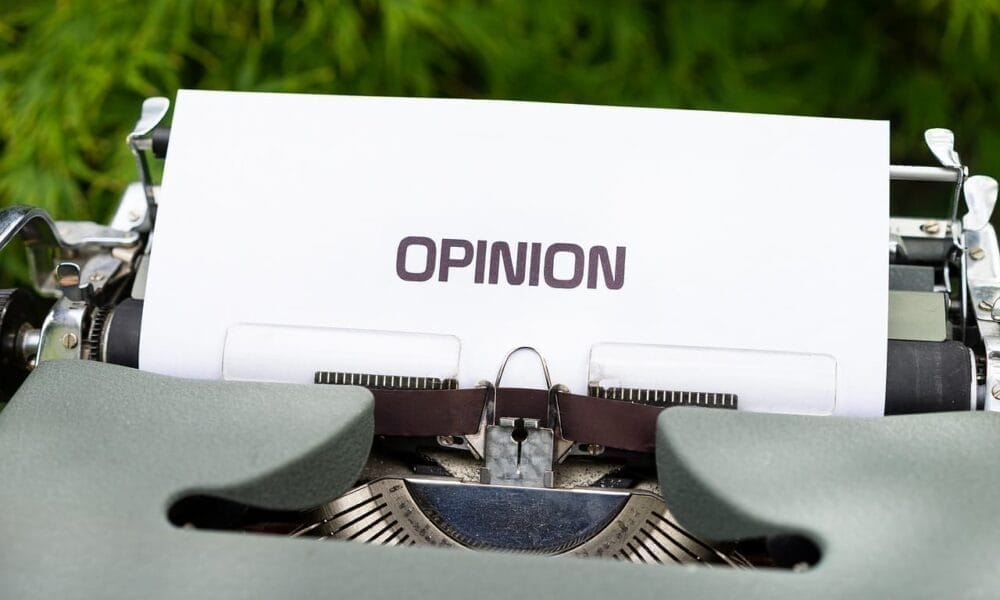 A close-up of a vintage typewriter with a sheet of white paper inserted displays the word "OPINION" in bold, uppercase letters. In the blurred background, green foliage adds contrast, hinting at controversial sports opinions waiting to be typed beneath.