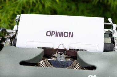 A close-up of a vintage typewriter with a sheet of white paper inserted displays the word "OPINION" in bold, uppercase letters. In the blurred background, green foliage adds contrast, hinting at controversial sports opinions waiting to be typed beneath.