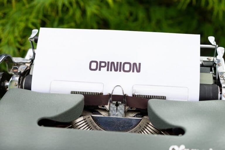 A close-up of a vintage typewriter with a sheet of white paper inserted displays the word "OPINION" in bold, uppercase letters. In the blurred background, green foliage adds contrast, hinting at controversial sports opinions waiting to be typed beneath.