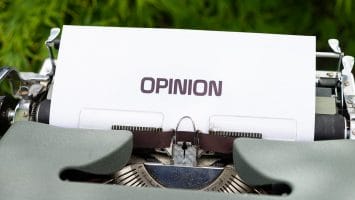 A close-up of a vintage typewriter with a sheet of white paper inserted displays the word "OPINION" in bold, uppercase letters. In the blurred background, green foliage adds contrast, hinting at controversial sports opinions waiting to be typed beneath.