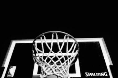 A basketball is caught in mid-air, reminiscent of 90s basketball glory, as it’s about to swoosh through the hoop. The hoop, mounted on a Spalding-branded backboard, stands out against the dark background, spotlighting this iconic moment.