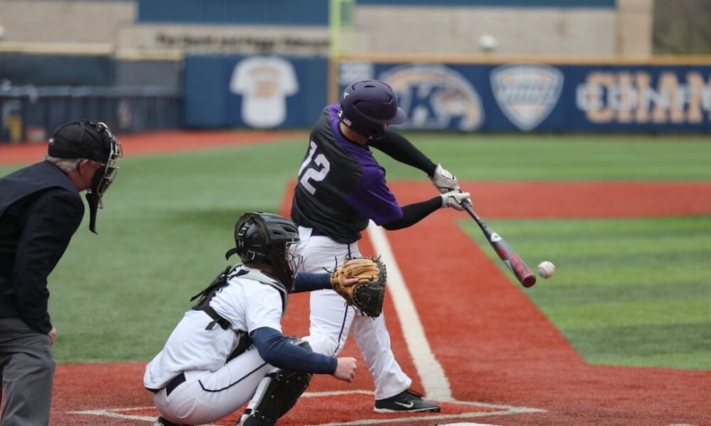 Exploring baseball, a player in a purple helmet and jersey, numbered 12, swings a bat at a pitched ball during the game. A catcher in white gear and an umpire stand behind him on the red and green field.