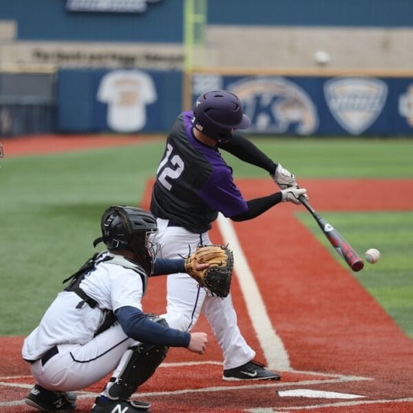 Exploring baseball, a player in a purple helmet and jersey, numbered 12, swings a bat at a pitched ball during the game. A catcher in white gear and an umpire stand behind him on the red and green field.