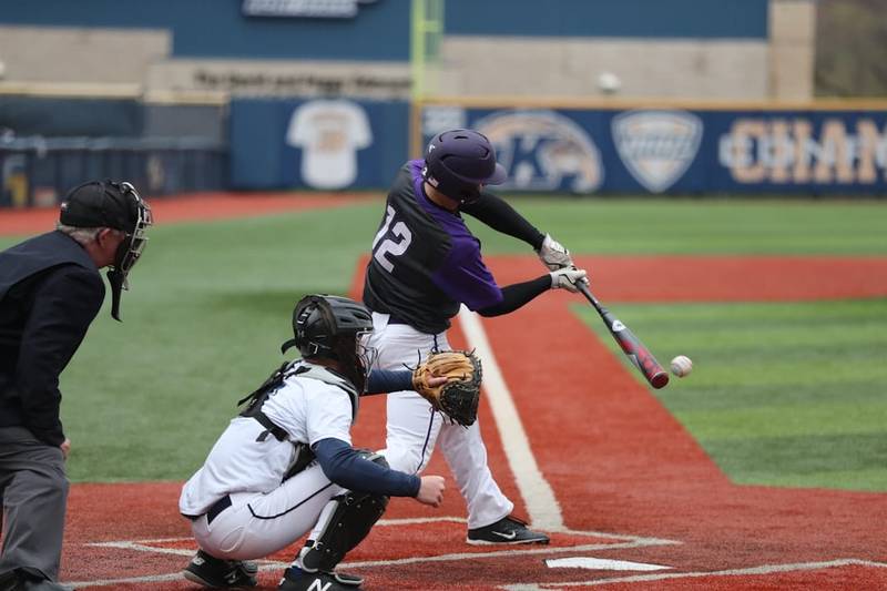 Exploring baseball, a player in a purple helmet and jersey, numbered 12, swings a bat at a pitched ball during the game. A catcher in white gear and an umpire stand behind him on the red and green field.