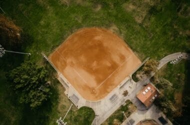 An aerial view of a baseball field showcases a pristine dirt infield and bases, perfect for scouts and data analysts alike. A small building nearby, possibly a dugout or storage area, stands amidst lush green grass, while floodlights tower overhead to illuminate every detail.