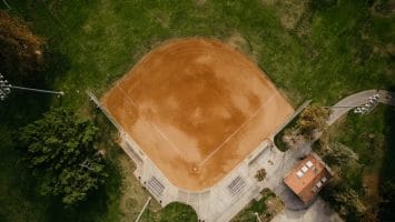 An aerial view of a baseball field showcases a pristine dirt infield and bases, perfect for scouts and data analysts alike. A small building nearby, possibly a dugout or storage area, stands amidst lush green grass, while floodlights tower overhead to illuminate every detail.