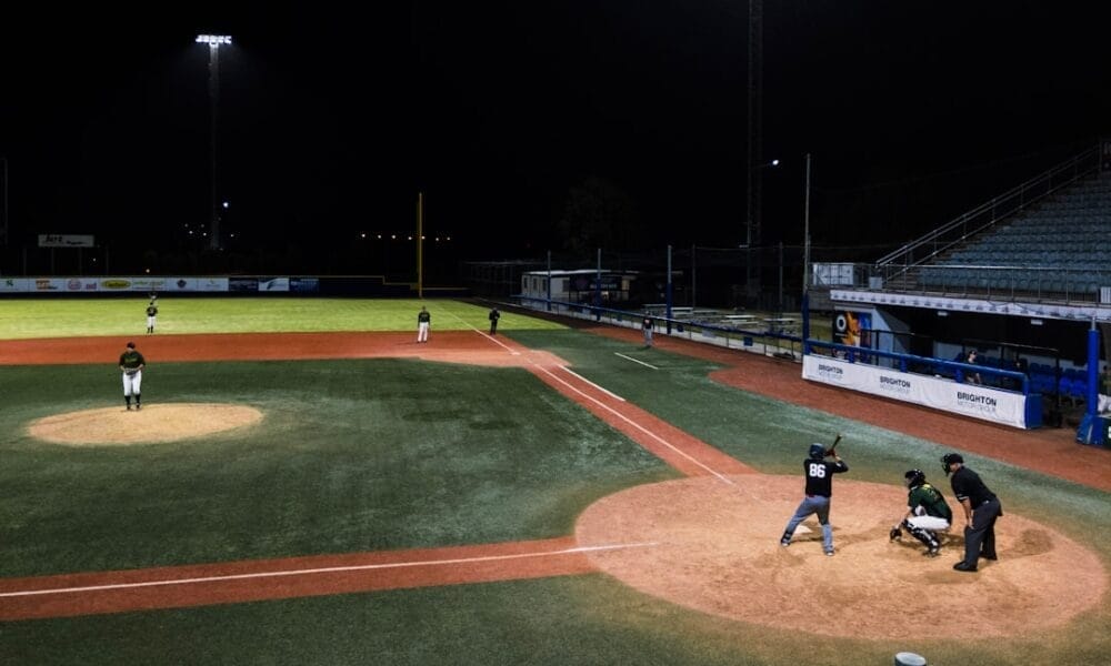 A traditional baseball night game unfolds under the lights, with a batter, catcher, and umpire at home plate. Players are perfectly positioned on the field as the classic sport comes alive under the night sky.