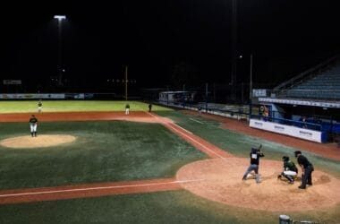 A traditional baseball night game unfolds under the lights, with a batter, catcher, and umpire at home plate. Players are perfectly positioned on the field as the classic sport comes alive under the night sky.