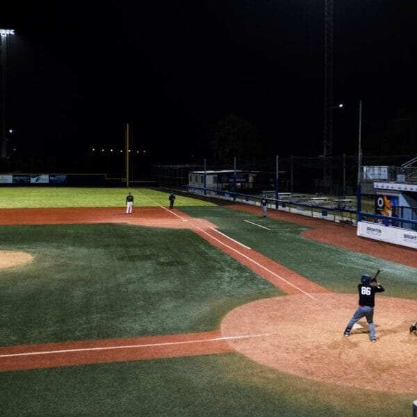 A traditional baseball night game unfolds under the lights, with a batter, catcher, and umpire at home plate. Players are perfectly positioned on the field as the classic sport comes alive under the night sky.
