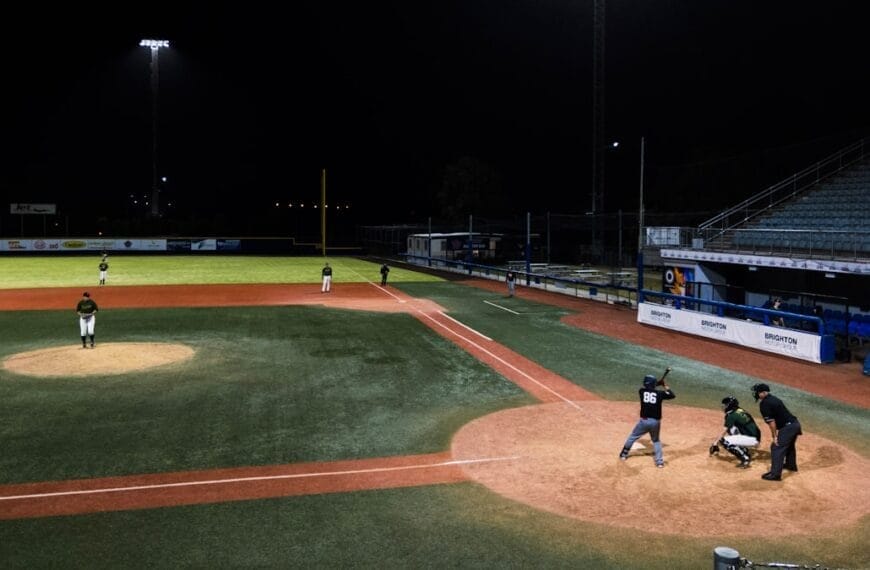 A traditional baseball night game unfolds under the lights, with a batter, catcher, and umpire at home plate. Players are perfectly positioned on the field as the classic sport comes alive under the night sky.