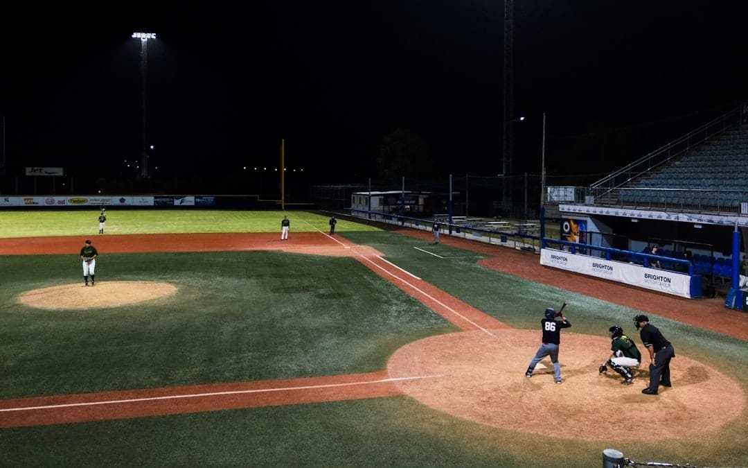 A traditional baseball night game unfolds under the lights, with a batter, catcher, and umpire at home plate. Players are perfectly positioned on the field as the classic sport comes alive under the night sky.
