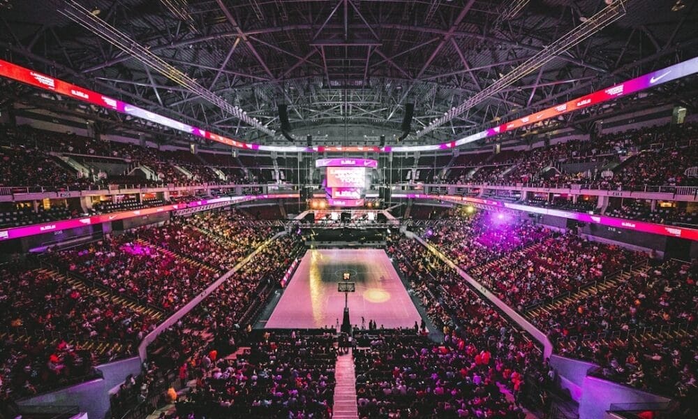 A large indoor basketball arena filled with spectators buzzes with NBA debates. The court is at the center, surrounded by tiered seating. Bright lights illuminate the space, and a scoreboard hangs above. The atmosphere is energetic and lively.