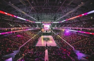 A large indoor basketball arena filled with spectators buzzes with NBA debates. The court is at the center, surrounded by tiered seating. Bright lights illuminate the space, and a scoreboard hangs above. The atmosphere is energetic and lively.