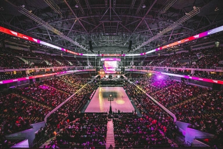 A large indoor basketball arena filled with spectators buzzes with NBA debates. The court is at the center, surrounded by tiered seating. Bright lights illuminate the space, and a scoreboard hangs above. The atmosphere is energetic and lively.