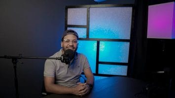 A person, likely a sports fan, wearing a cap and glasses sits at a table with a microphone in front of them. The background features a lit panel with a blue glow and a lamp casting purple and pink lighting, creating the perfect ambiance for recording spirited podcasts.