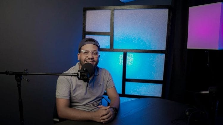 A person, likely a sports fan, wearing a cap and glasses sits at a table with a microphone in front of them. The background features a lit panel with a blue glow and a lamp casting purple and pink lighting, creating the perfect ambiance for recording spirited podcasts.