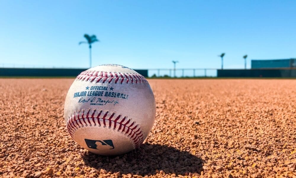 A baseball showcasing its stitching and text rests on a dirt field under a clear blue sky. The blurred background reveals a fence and two palm trees, an idyllic setting for contemplating baseball strategies.