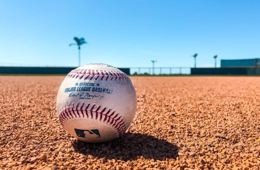A baseball showcasing its stitching and text rests on a dirt field under a clear blue sky. The blurred background reveals a fence and two palm trees, an idyllic setting for contemplating baseball strategies.